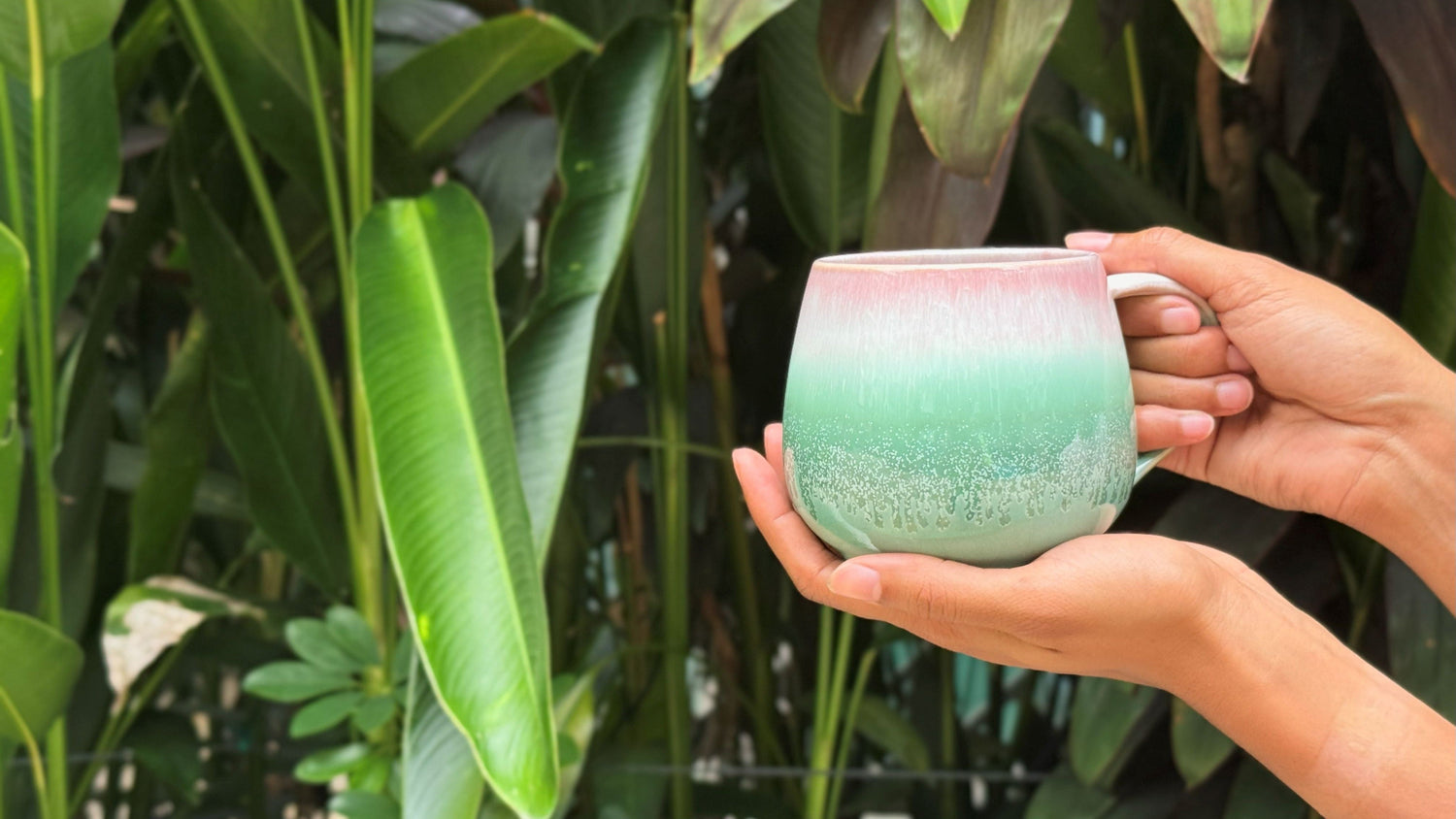 Person holding a green ceramic mug with plants in the background